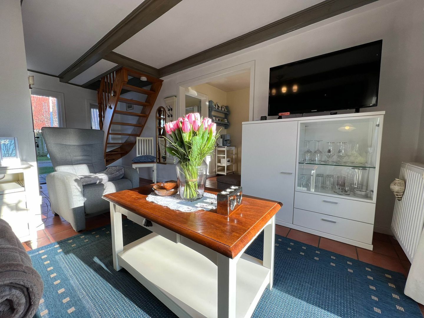 A cozy living room with a wooden coffee table, a grey armchair, and a vase of pink tulips. There's a white cabinet with glassware and a flat-screen TV mounted above it.