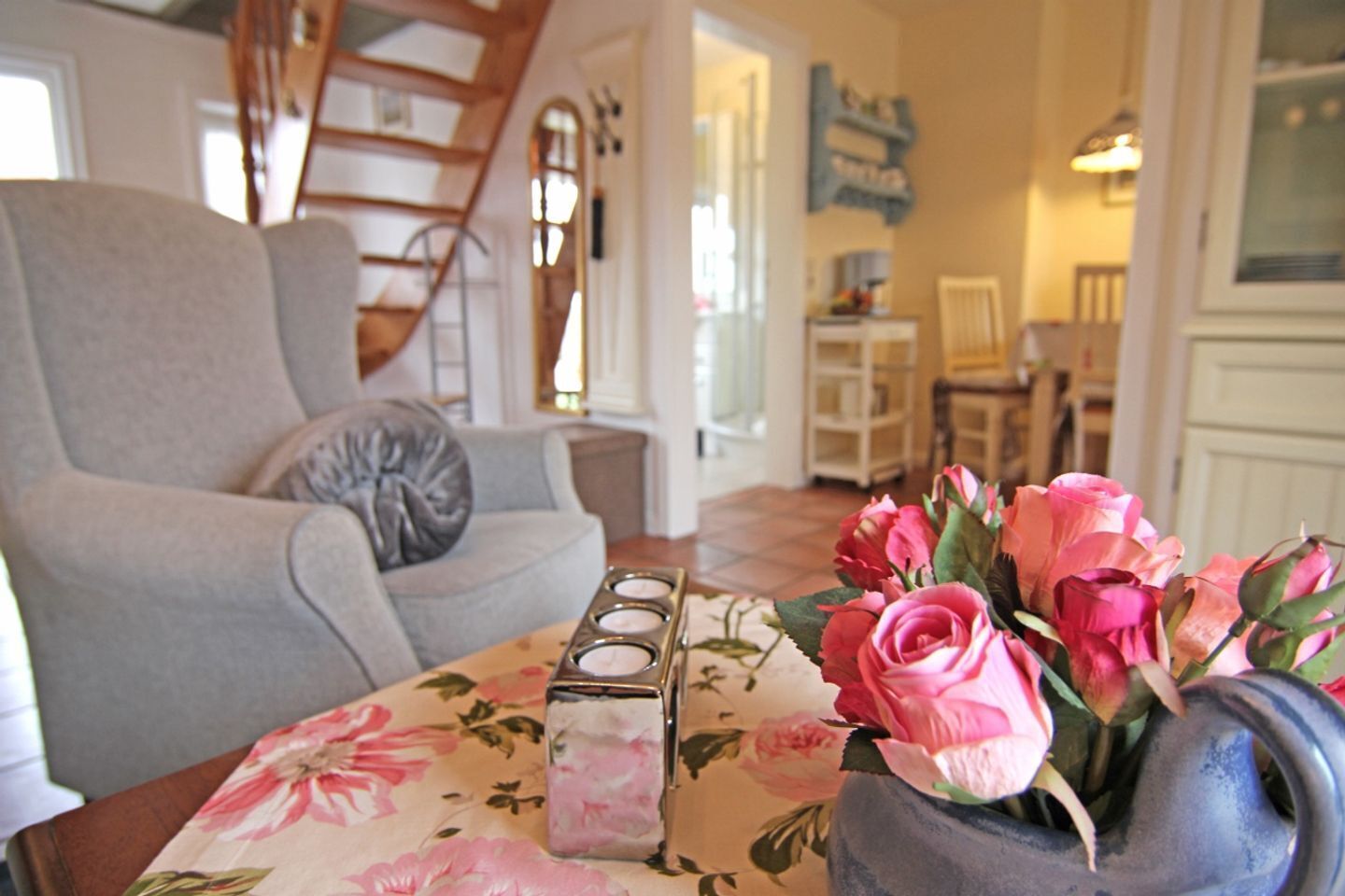 A living room with a grey sofa, a floral tablecloth, a vase of pink roses, and a set of candles.
