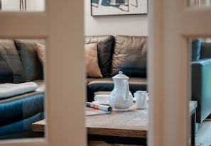 Ein gemütliches Wohnzimmer mit einer Ledersofa, einem Holztisch mit einem weißen Teekessel, Tassen und einer aufgerollten Zeitschrift.