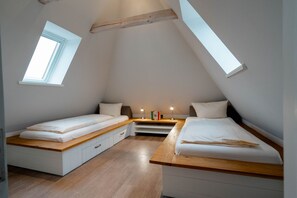 Un dormitorio en ático pequeño y triangular con dos camas individuales, una mesita de noche con lámpara y un estante pequeño con libros.