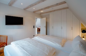 A bedroom with two beds, a TV mounted on the wall, wooden beams on the ceiling, and a wardrobe with sliding doors.