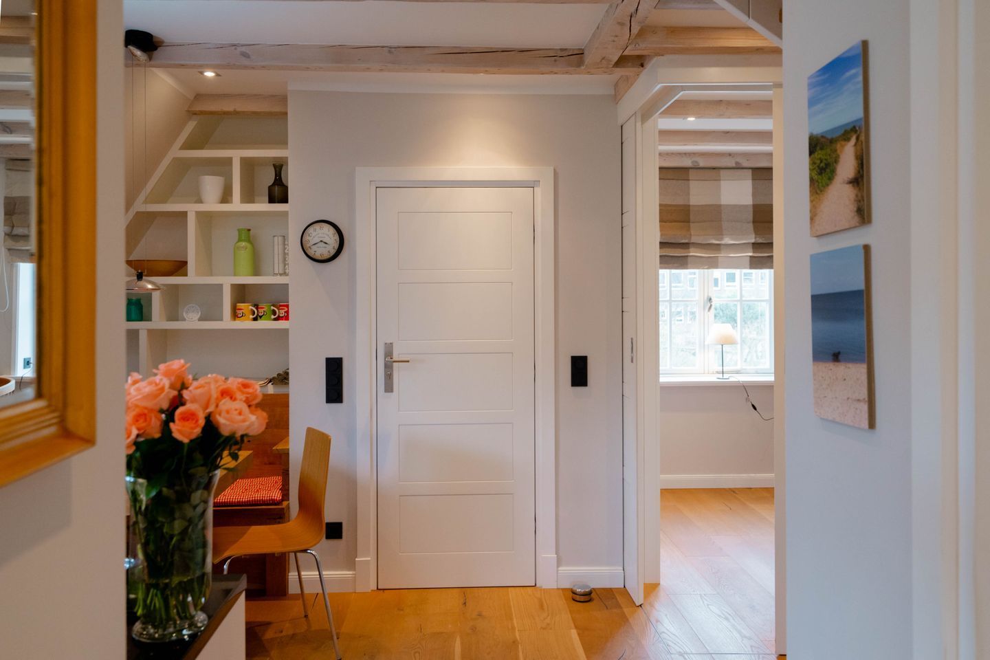 Un couloir avec une porte blanche, des étagères en bois, une horloge et une fenêtre donnant sur l’extérieur.