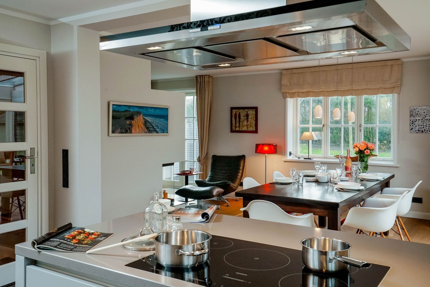A modern kitchen with an induction cooktop, a dining area with a table set for two, and a view of the outdoors through large windows.