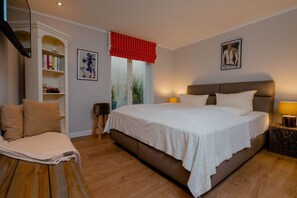 A bedroom with a large bed, a bookshelf, a small stool, and a window with a red valance.