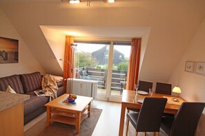 A cozy living room with a sofa, coffee table, and dining table with chairs. A large window offers a view of the outdoors. - Henning 1 floor - Moin Moin and welcome to the cozy 3-room (Wenningstedt-Braderup (Sylt))