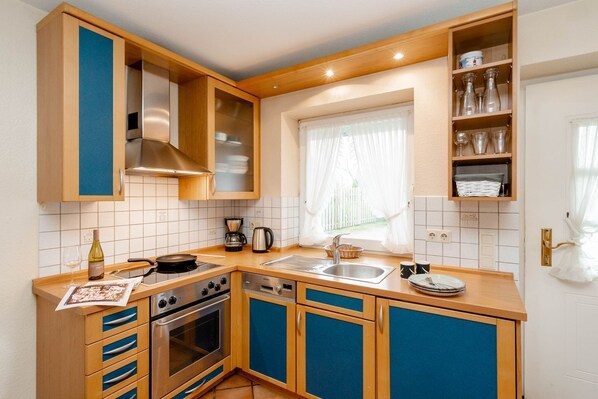 A compact kitchen with wooden cabinets, a built-in oven, a sink, and a window with curtains. - Godewind ground floor - Moin Moin and welcome to the ground floor 3-room (Sylt-Ost)