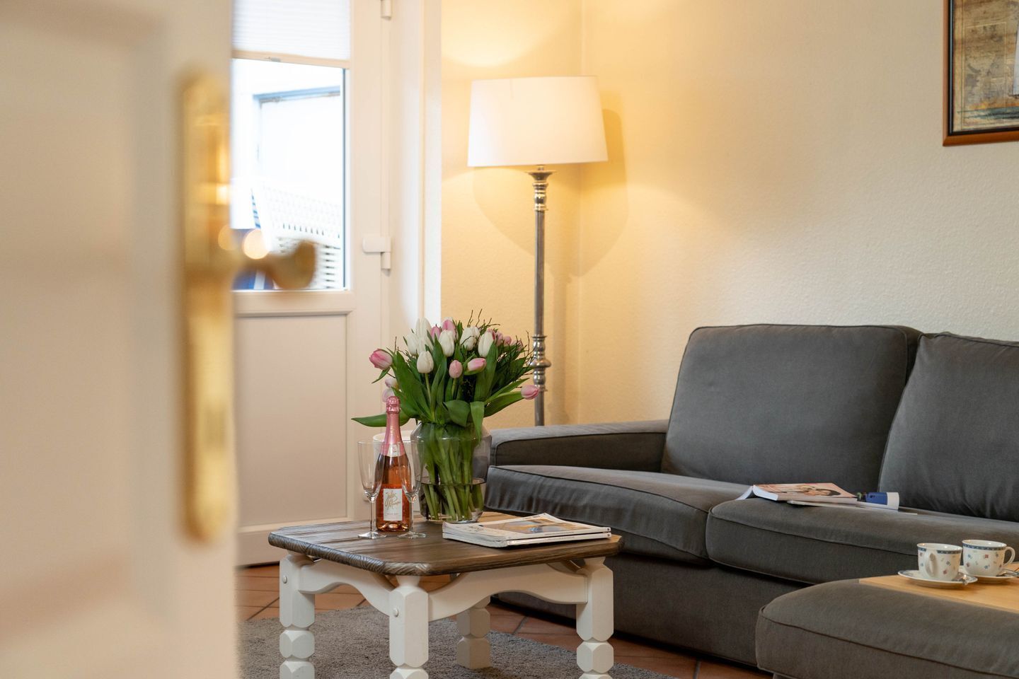 A living room with a grey sofa, a small table with a vase of flowers, a lamp, and a bottle of wine.