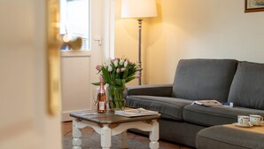 A living room with a grey sofa, a small table with a vase of flowers, a lamp, and a bottle of wine. - Godewind ground floor - Moin Moin and welcome to the ground floor 3-room (Sylt-Ost)