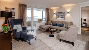 Een moderne woonkamer met een grijze fauteuil, een beige bank en een salontafel. Er is een raam met gordijnen, een lamp en een ingelijst kunstwerk aan de muur.