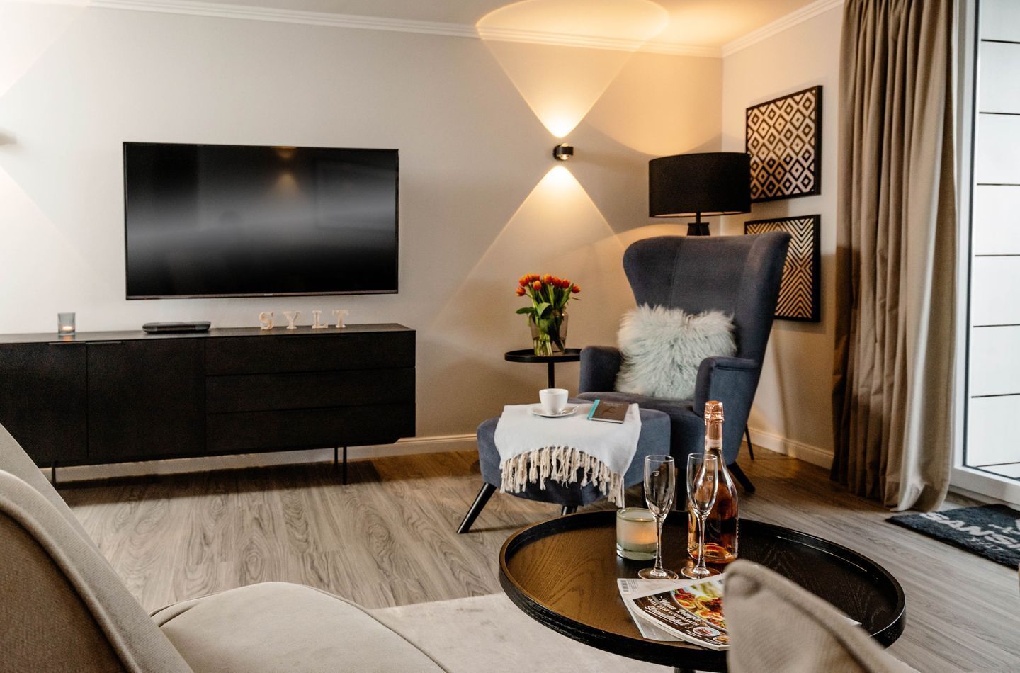 A modern living room with a flat-screen TV, a dark armchair, a round coffee table with drinks, and a small side table with a vase of flowers.