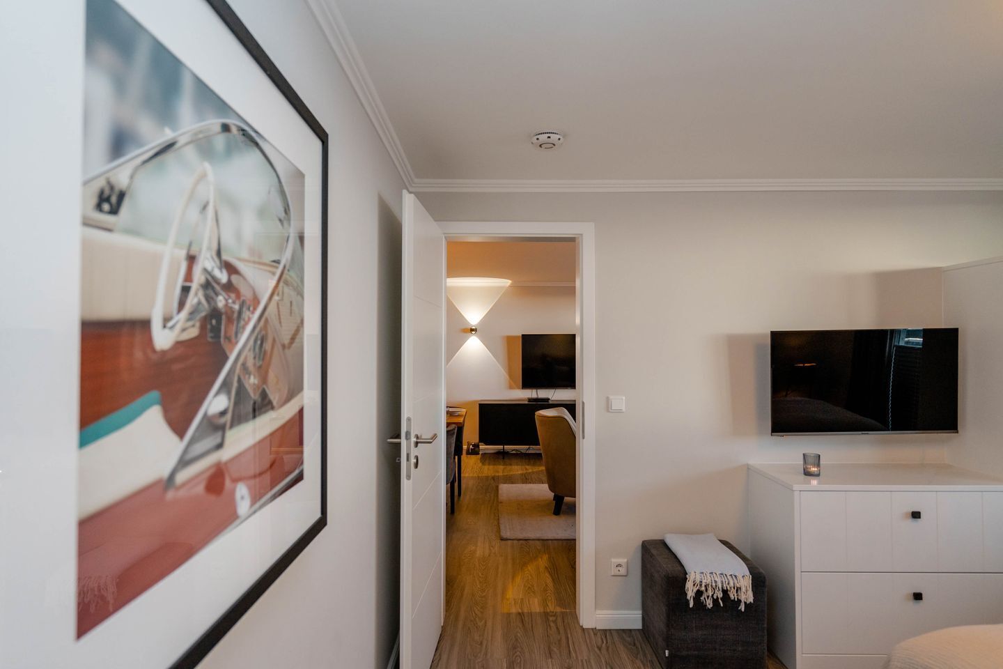 A modern hotel room with a large framed picture, a flat-screen TV mounted on the wall, a small bedside table, and a wooden floor.