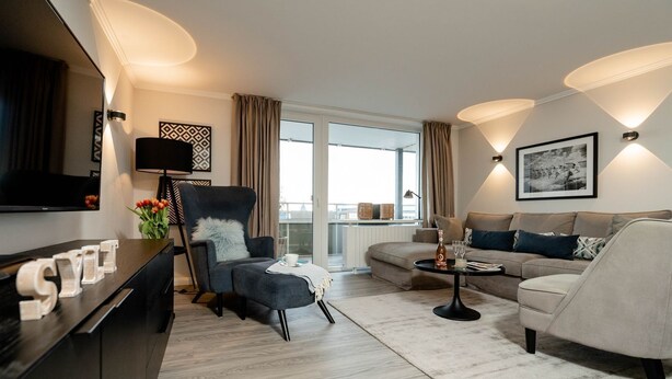 Een moderne hotelkamer met een bank, fauteuil en een eettafel. Er is een televisie aan de muur bevestigd, en een raam met gordijnen.