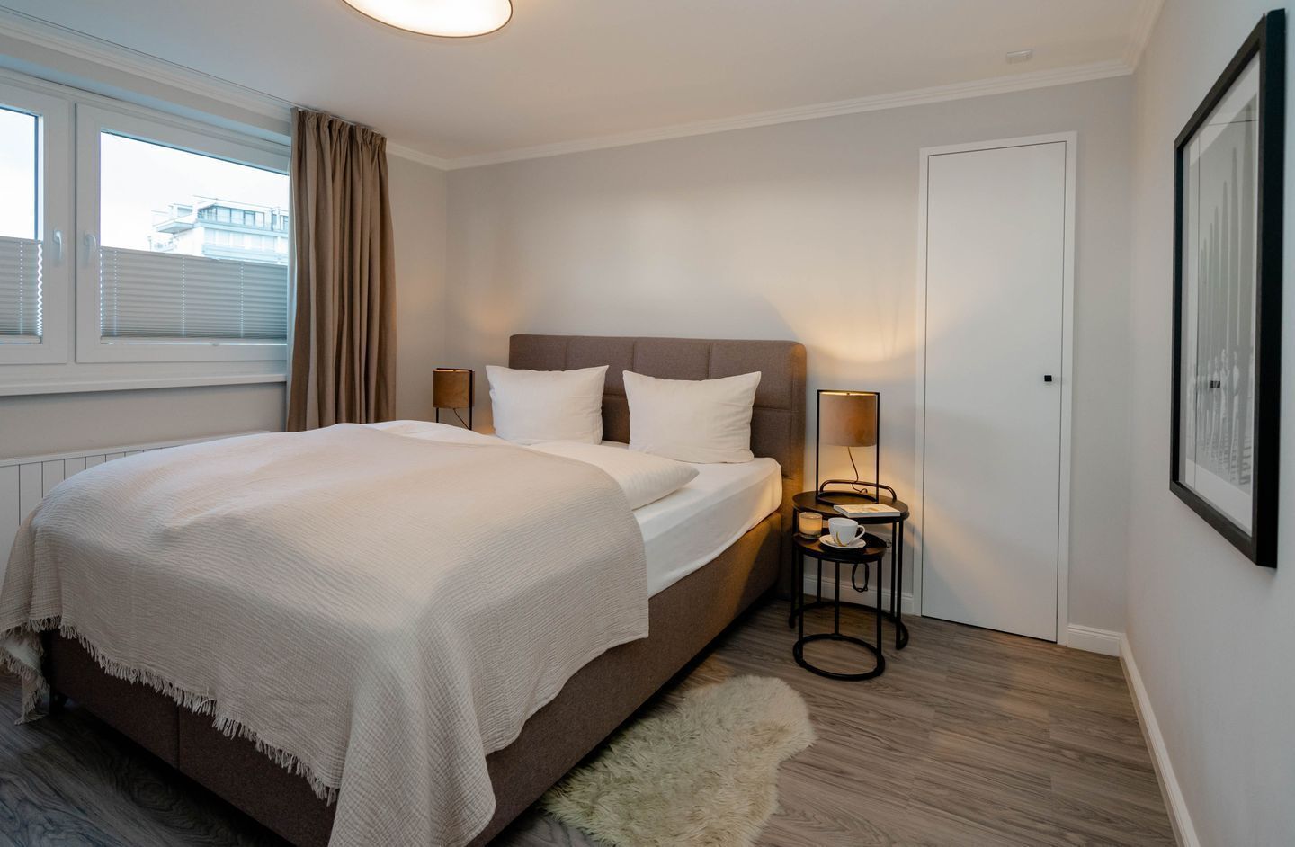 A hotel room with a bed, bedside lamps, a small table with a cup and saucer, a framed picture on the wall, and a window with blinds.