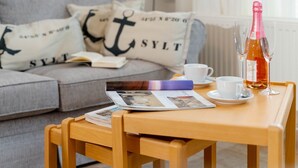 Una mesa de madera con una botella de vino, dos copas, una taza y una revista. Un sofá gris con almohadones de ancla se ve al fondo.