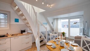 Una cocina moderna con encimera de madera, armarios blancos y una estufa integrada. Hay un comedor con una mesa redonda de madera preparada para cuatro, rodeada de sillas blancas. Una escalera con barandillas blancas conduce a un nivel superior.