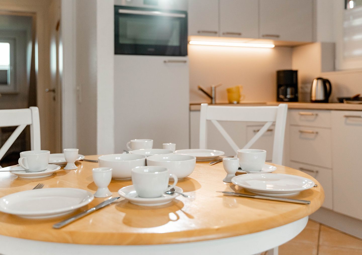 A dining table set with white plates, cups, and cutlery in a modern kitchen with white cabinets and a microwave.