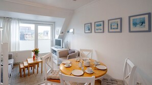 Un comedor con una mesa redonda preparada para cuatro, sillas blancas, un televisor sobre un mueble y una ventana con cortinas.