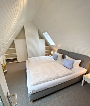 A bedroom with a sloped ceiling, a bed with white bedding, a nightstand, and a small table with a lamp.