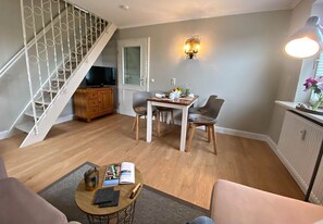 A living room with a staircase, a dining table with chairs, a television, and a wall-mounted light fixture.