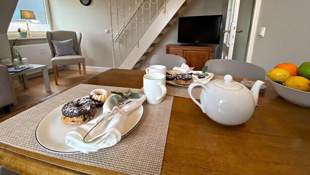 Un coin repas avec une table dressée pour deux personnes, agrémentée d’une assiette de pâtisseries, d’une théière et d’un saladier de fruits.