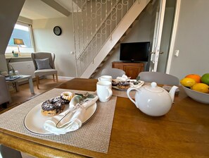 Un coin repas avec une table dressée pour deux personnes, agrémentée d’une assiette de pâtisseries, d’une théière et d’un saladier de fruits.