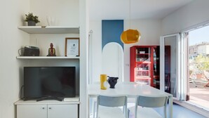 Una cuina moderna amb televisió de pantalla plana, armaris blancs, una taula de dinar amb caires, una prestatgeria vermella i una finestra gran.