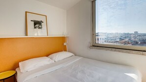 Una habitació d'hotel amb un llit, una finestra que dona a la ciutat, un quadre encimbellat a la paret i una taula petita.