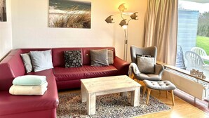 Een woonkamer met een rode bank, een grijze fauteuil, een houten salontafel en een kamerbreed tapijt. Er is een groot raam met gordijnen, een wandlamp en een ingelijste foto boven de bank.