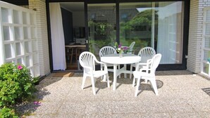 Een terras met een ronde tafel en vier witte stoelen, een kleine tuin met roze bloemen, en een schuifdeur naar een binnenruimte met een keuken en eettafel.