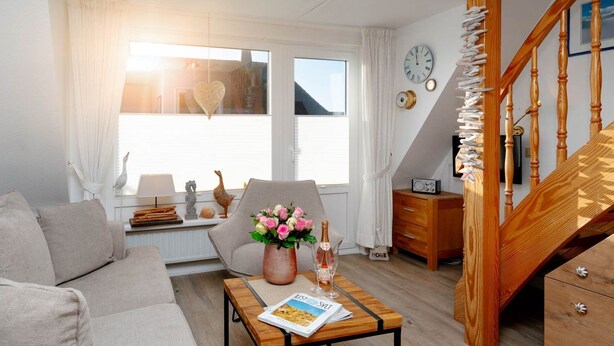 A cozy living room with a sofa, a coffee table with a magazine, a chair, a vase of flowers, and a staircase with wooden railings.