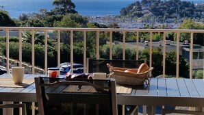 Een balkon met een tafel, stoelen, een mand en uitzicht op zee en gebouwen.