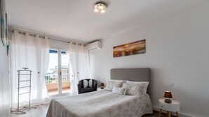 Een slaapkamer met een bed, een stoel, een klein tafeltje met een lamp en een balkon met een hekwerk.