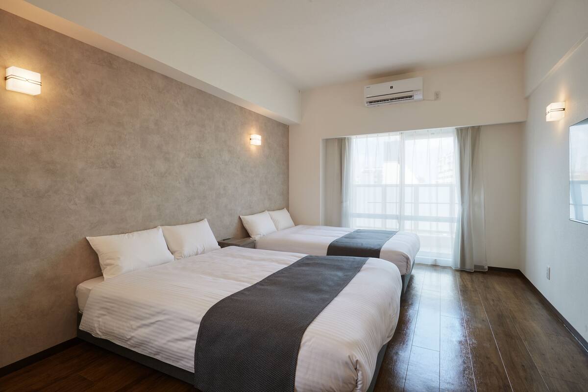 Quarto de hotel com duas camas, uma janela grande com cortinas e um aparelho de ar condicionado na parede.
