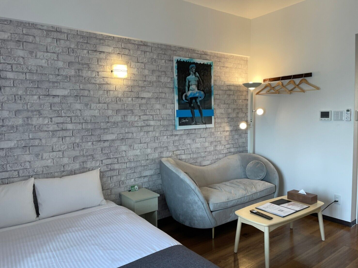 A modern hotel room with a brick-patterned wall, a bed with white linens, a grey armchair, a small bedside table with a lamp, and a poster on the wall.