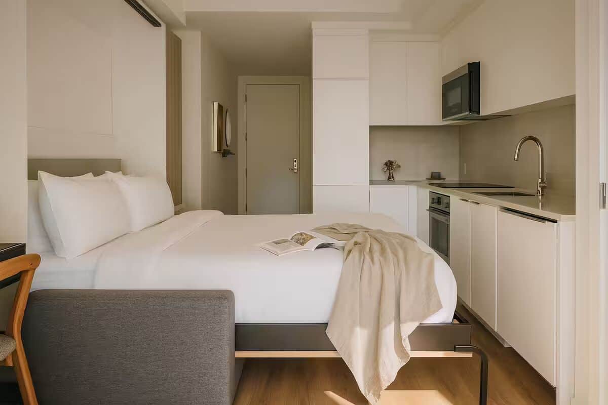 Una habitación de hotel moderna con cama, un pequeño escritorio, una silla, una kitchenette con microondas y una puerta que da a otra habitación.
