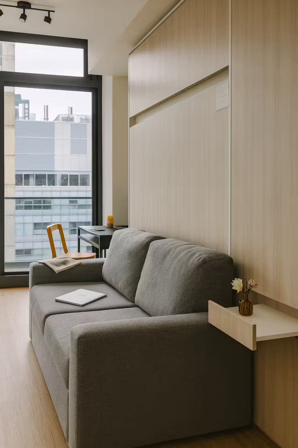 Una habitación moderna con un sofá gris, una mesita, una silla, un ventanal con vistas a la ciudad y una estantería de madera con un florero.