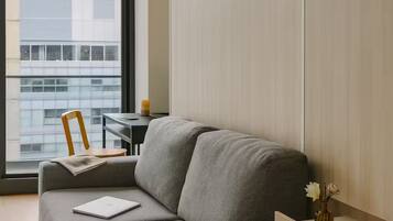 Una habitación moderna con un sofá gris, una mesita, una silla, un ventanal con vistas a la ciudad y una estantería de madera con un florero.