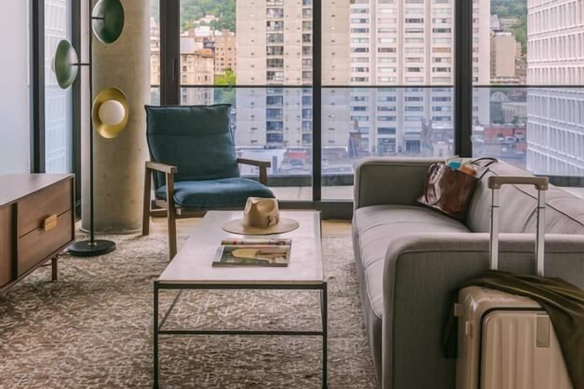 Una habitación de hotel moderna con un ventanal grande, un sofá, una mesita de café, una silla y una maleta.