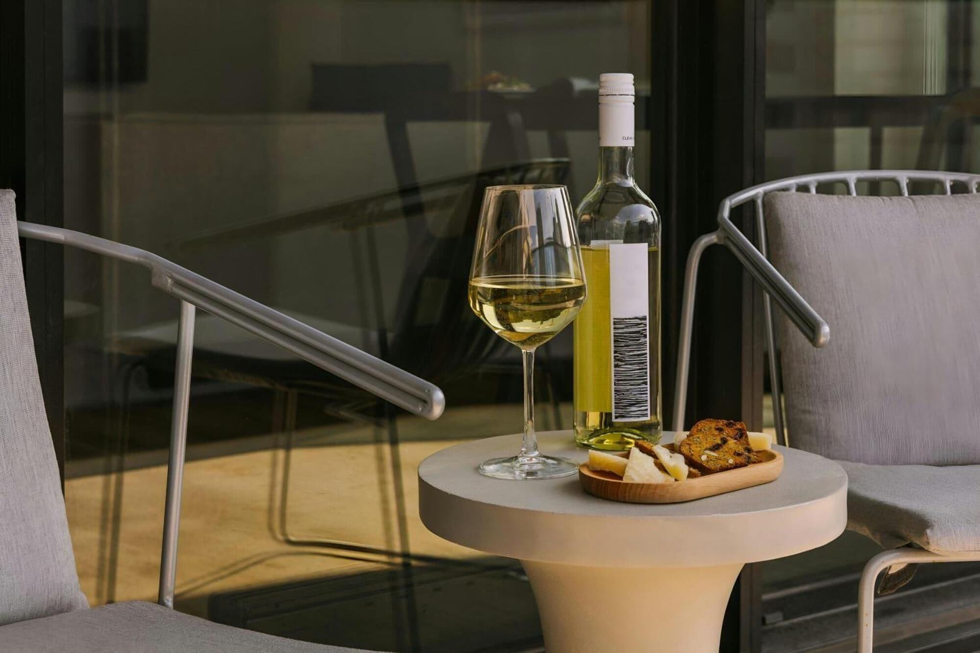 A round table with a bottle of white wine, a glass, and a plate of cheese and crackers on a balcony.