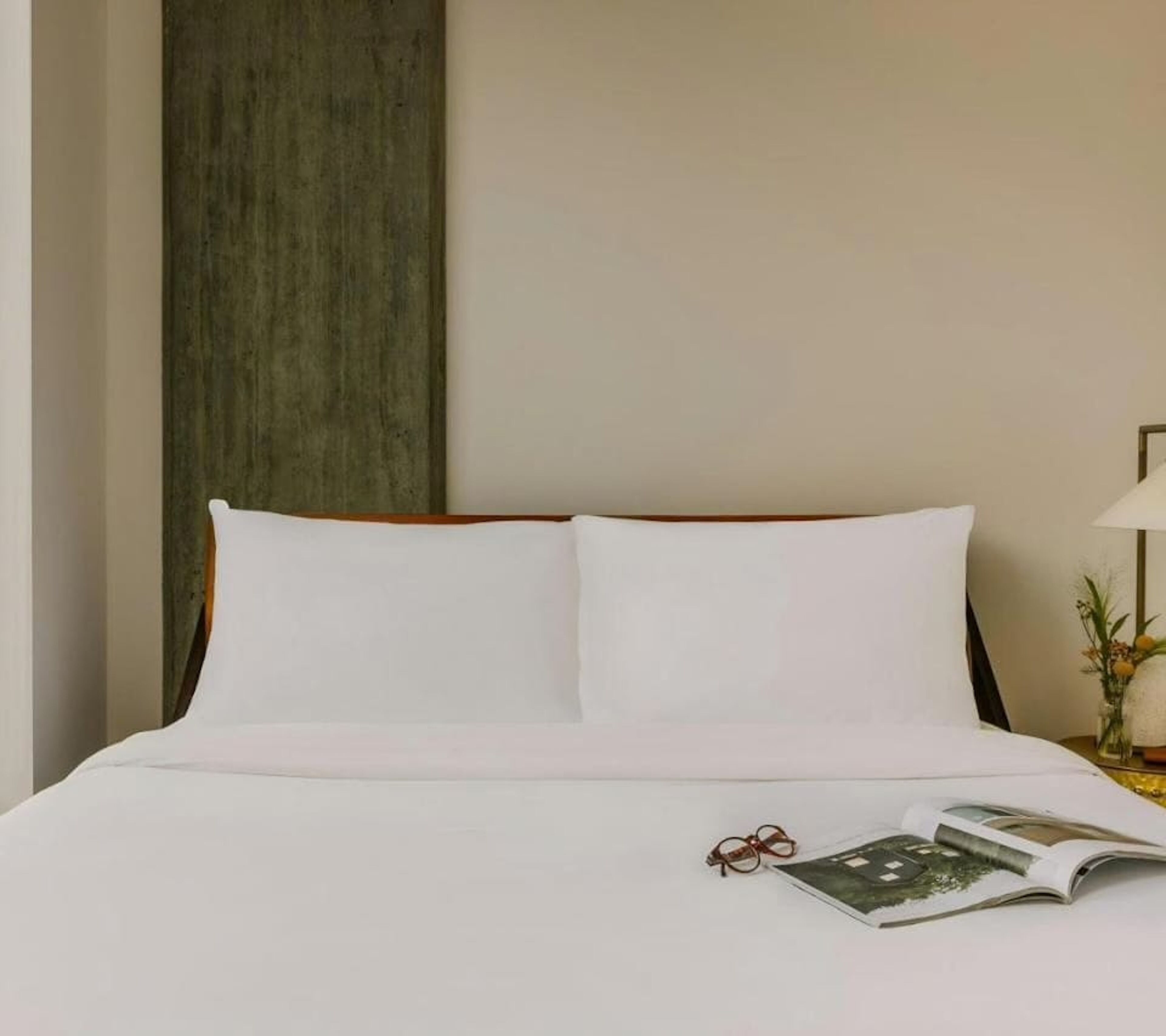 Una cama bien hecha con sábanas blancas, un par de gafas y una revista abierta sobre la cama.