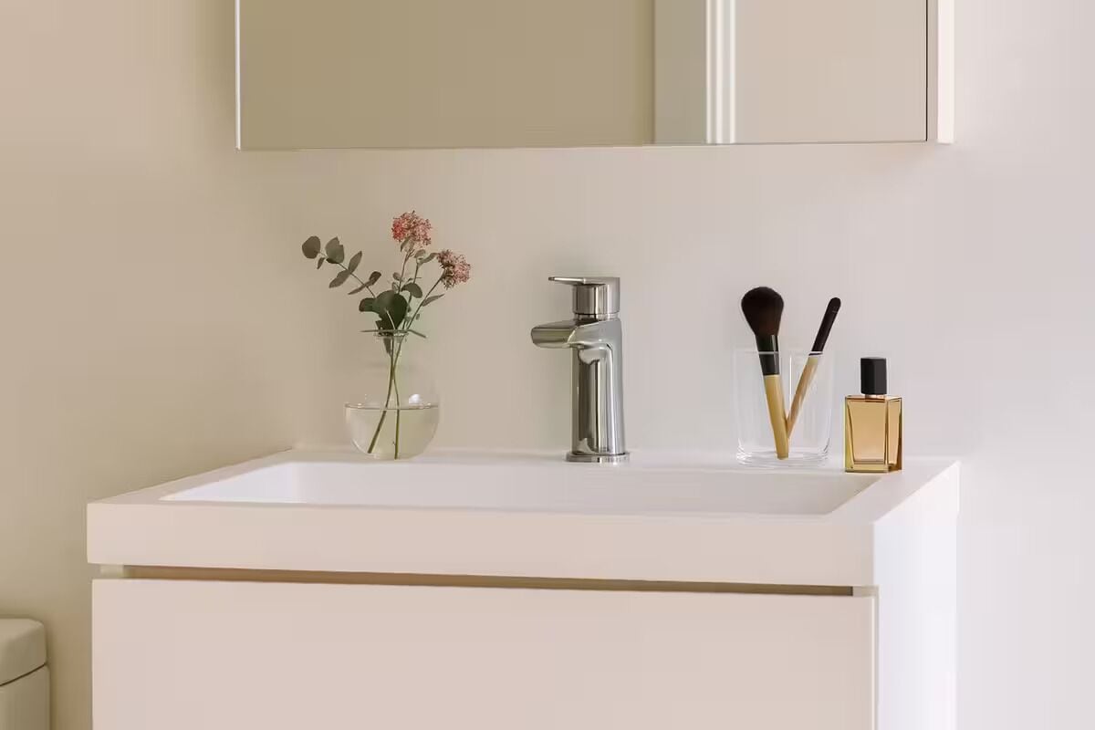 A bathroom with a white sink, a mirror above it, a vase with flowers, and a bottle of perfume.
