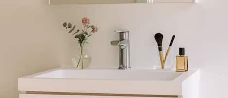 A bathroom with a white sink, a mirror above it, a vase with flowers, and a bottle of perfume.