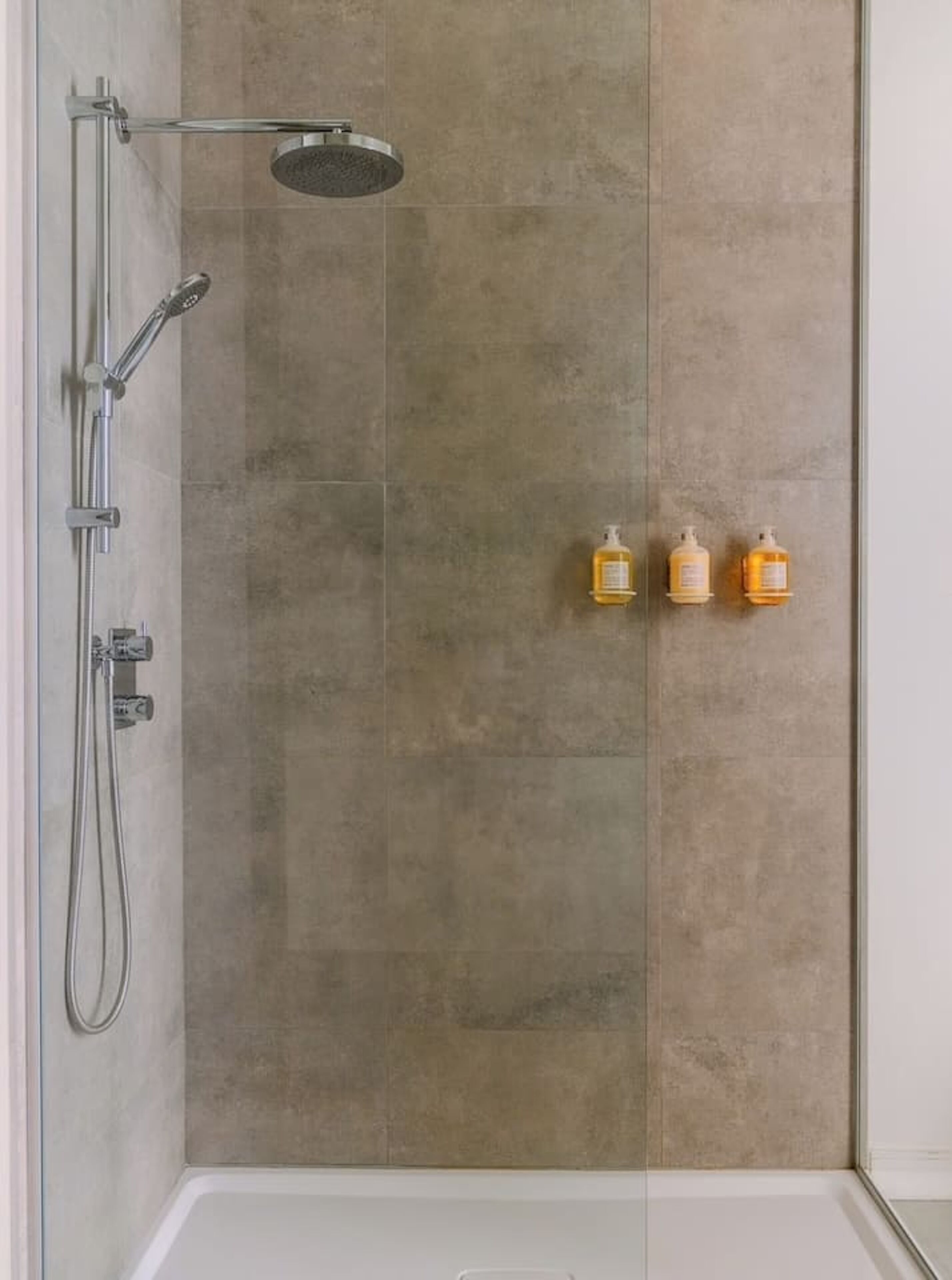 A shower with a glass door, a handheld showerhead, and two bottles of shampoo on the wall.