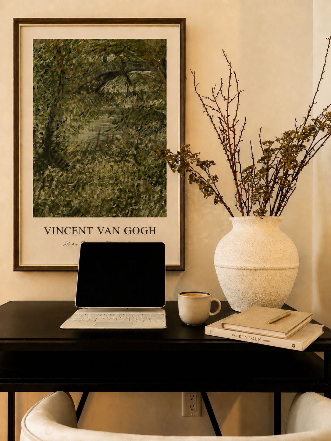 Ein gerahmtes Kunstwerk von Vincent van Goghs ‚River of Trees‘ an der Wand, ein Laptop auf einem Schreibtisch, eine Vase mit Ästen, eine Tasse Kaffee und ein Buch mit dem Titel ‚The Kinfolk Home‘.