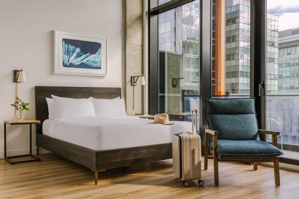 Habitación de hotel con cama, una silla, una maleta, una lámpara y un cuadro en la pared.