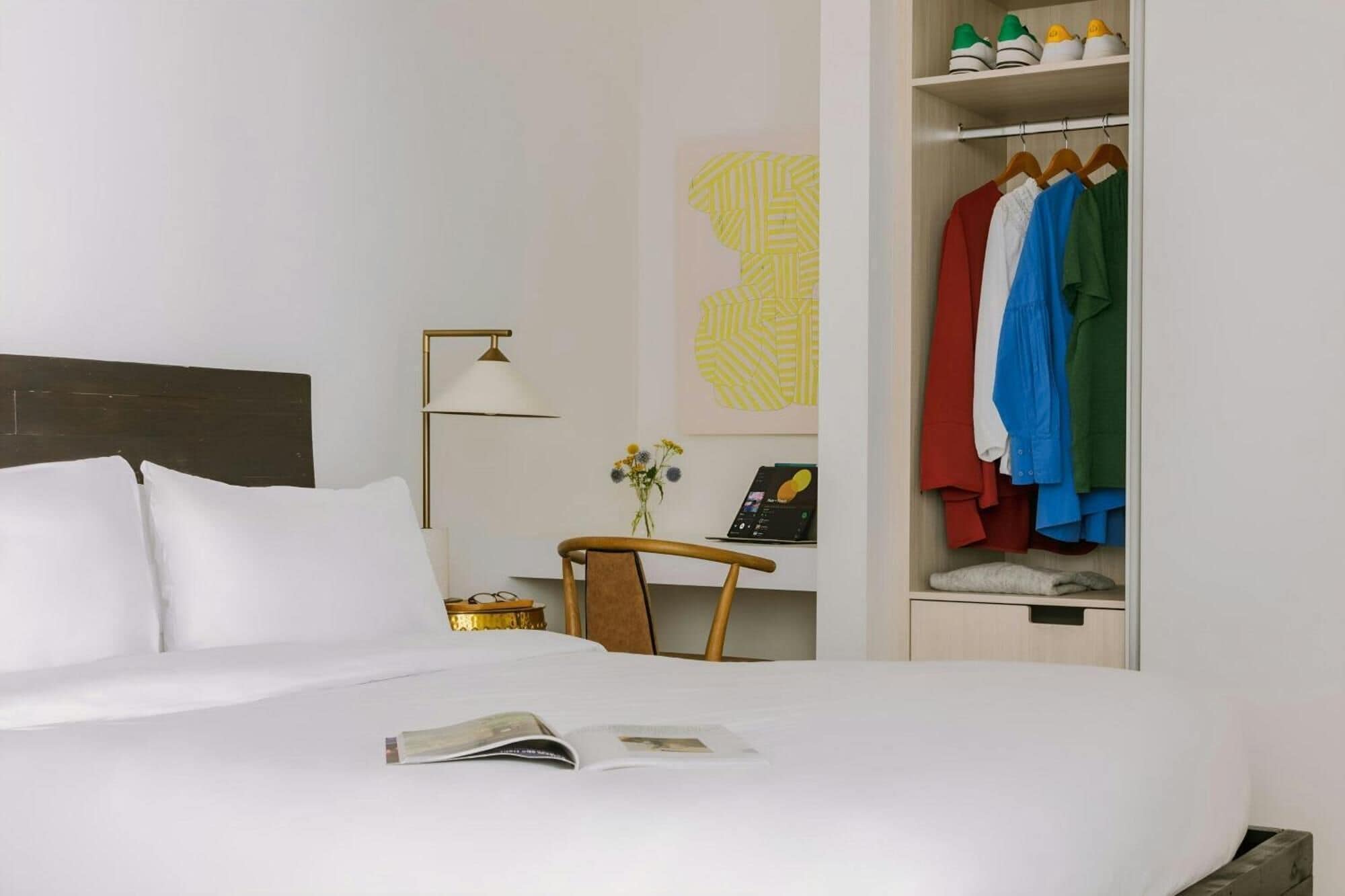 A hotel room with a bed, a desk with a lamp, a chair, a small table with a vase, and a wardrobe with clothes.