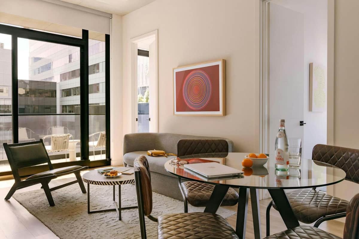 Una sala de estar moderna con una mesa de vidrio, sillas de cuero, un sofá y un amplio ventanal con vistas a un edificio.