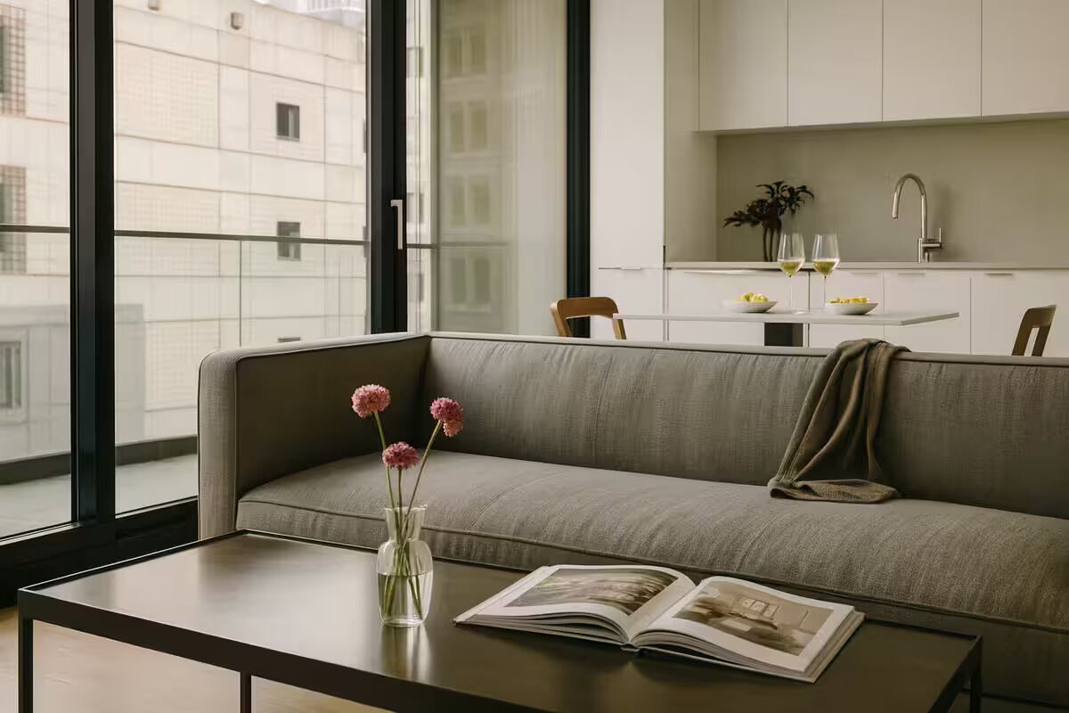 Un salon moderne avec un canapé, une table basse et un livre. Une fenêtre offre une vue sur les bâtiments à l’extérieur.