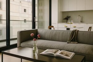 Un salon moderne avec un canapé, une table basse et un livre. Une fenêtre offre une vue sur les bâtiments à l’extérieur.