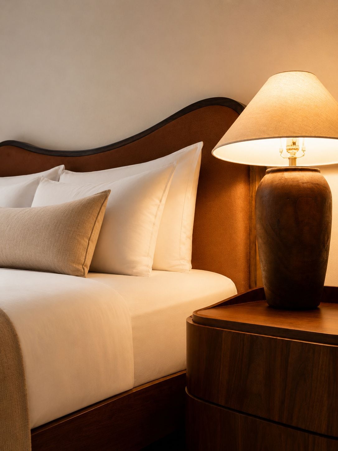 A neatly made bed with white and beige bedding, a wooden headboard, and a round wooden nightstand with a lamp.
